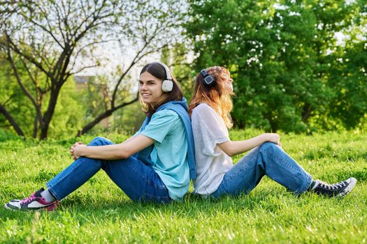 Two friends guy girl listen to music podcast, in wireless headphones, sitting on lawn on grass. Enjoying friendship, music, nature. Youth, lifestyle, young people concept
