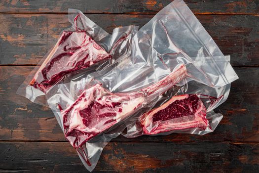 Raw uncooked beef steaks in vacuum packed air sealed sous vide bag set, tomahawk, t bone and club steak cuts, on old dark wooden table background, top view flat lay