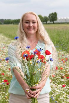 a beautiful middle-aged blonde woman stands among a flowering field of poppy, daisies, Cornflowers, and holds a bouquet of wild flowers and laughs. High quality photo