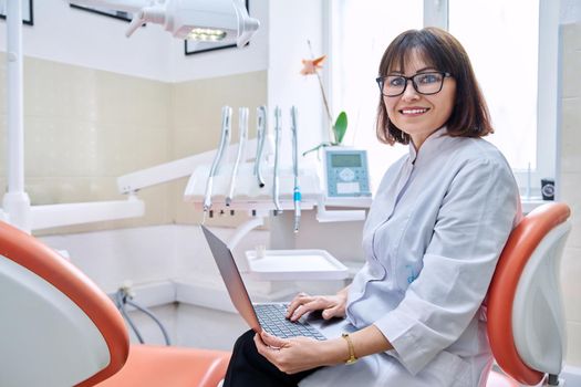 Portrait of dentist doctor sitting in office with laptop in her hands looking at camera. Mature smiling female nurse in dental clinic. Dentistry, medicine, health care, profession, stomatology concept
