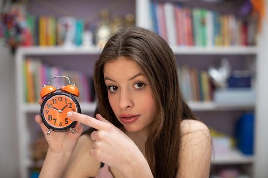 A young girl points her finger at an old orange alarm clock. A young brunette with long hair announces that the appointed time will soon pass.