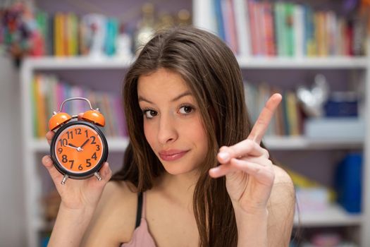 A young teenage girl warns about being behind schedule and shows her alarm clock. A young brunette raises her finger up giving a warning for not being on time.