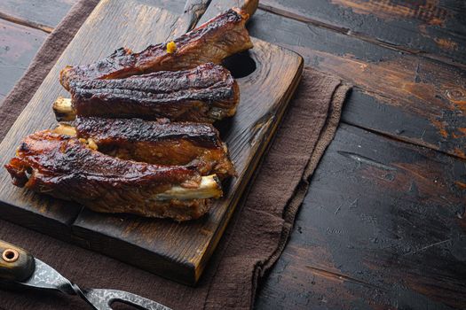 Grilled barbecue pork ribs set, on wooden serving board, with barbeque knife and meat fork, on old dark wooden table background
