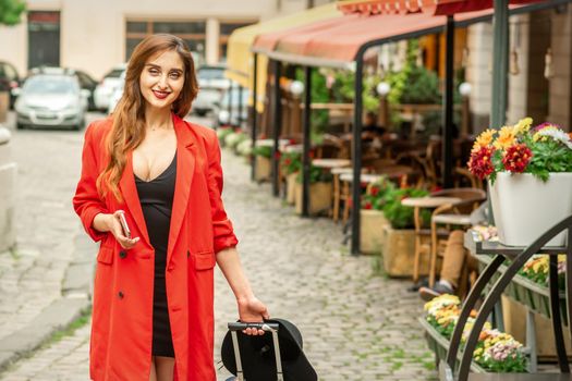 Beautiful fashionable young brunette woman walking at city street with travel suitcase and smartphone