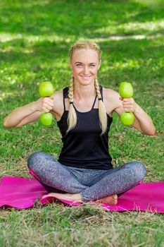 Young woman is training with apple dumbbells sitting on the grass in the park