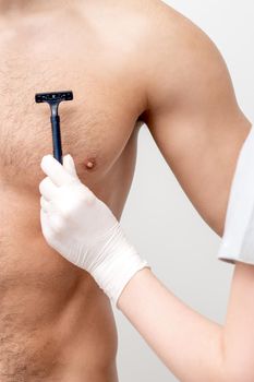 Hand of female beautician shaves chest of young man by razor. Concept of hair removal