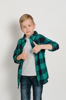 Portrait of cute stylish blond boy kid 7 years old in checked shirt and jeans showing thumb up