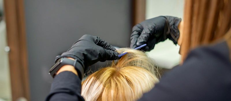 Female hairdresser makes hairstyle for young blonde woman wearing gloves in hair salon