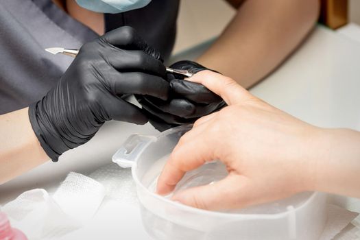 Manicurist removing cuticle from female nails by metal pusher during soaking fingernails in the bath at nail salon