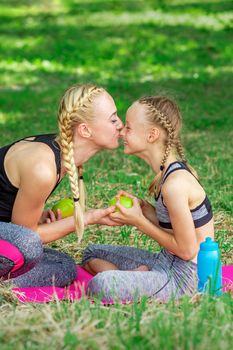 Mother kissing her daughter in nose giving green apple sitting on the grass in the park