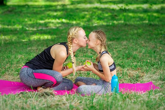 Mother kissing her daughter in nose giving green apple sitting on the grass in the park