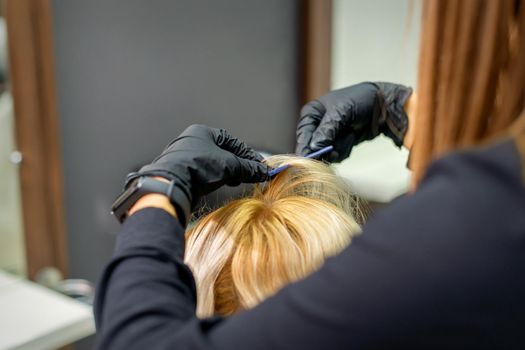 Female hairdresser makes hairstyle for young blonde woman wearing gloves in hair salon