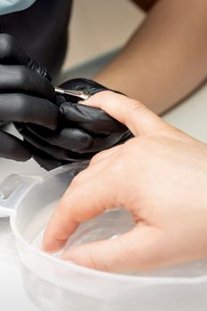 Manicurist removing cuticle from female nails by metal pusher during soaking fingernails in the bath at nail salon