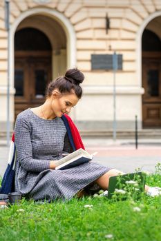 Young caucasian student woman reading the book sitting on the grass near university on the street