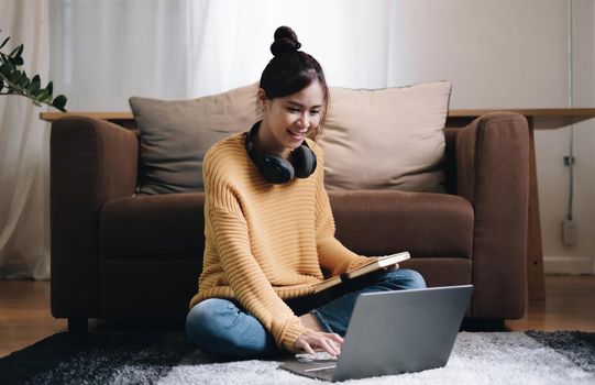 Smiling young woman working on laptop sitting on the floor in front of the sofa wearing headphones at home.