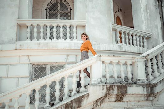 A middle-aged woman looks like a good blonde with curly beautiful hair and makeup on the background of the building. She is wearing a yellow sweater