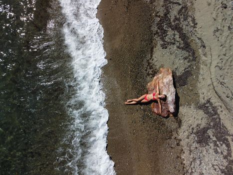 Young sensual blonde woman sitting on the rock near water at sea and enjoys the sea waves on background of two volcanic rocks, like in Iceland. Dreams holidays and weekend vacation in summer time