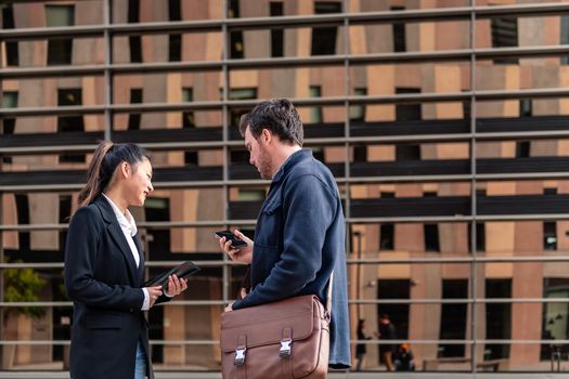 young business couple with mobile phone and tablet talking next to office building in the financial district, concept of entrepreneurship and business, copy space for text