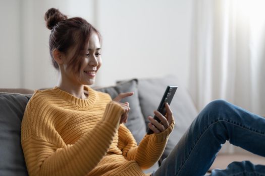 Happy asian teen girl checking social media holding smartphone at home. Smiling young woman using mobile phone app playing game, shopping online, ordering delivery relax on sofa.