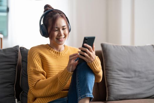 Freelance asian woman with mobile phone listening music in headphones and relax at home. Happy girl sitting on couch in living room