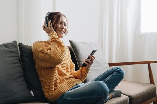 Freelance asian woman with mobile phone listening music in headphones and relax at home. Happy girl sitting on couch in living room