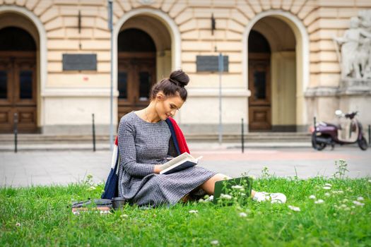 Young caucasian student woman reading the book sitting on the grass near university on the street