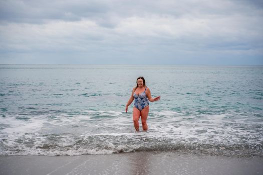 A plump woman in a bathing suit enters the water during the surf. Alone on the beach, Gray sky in the clouds, swimming in winter