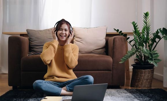 smiling girl looking at the camera Next to the laptop sit on the floor in front of the sofa and put on headphones to listen to music at home..