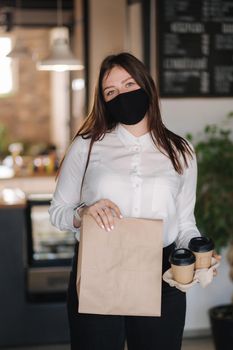 Cashier in face mask holding package with food and two cups of coffee. Contactless payments concept at cafe. Take away food during qurantine.