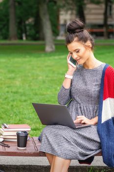 Student young woman is talking by smartphone using laptop on the bench in the park