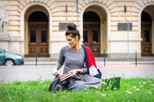 Young adult female student puts books to backpack sitting on grass near university building