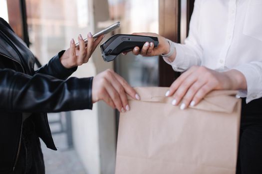 Closeup of female standing outdoors by cafe and paying with smartphone during Covid-19 pandemic. Cashier hand holding credit card reader machine while client holding phone for NFC payment. Focus on hand with package.