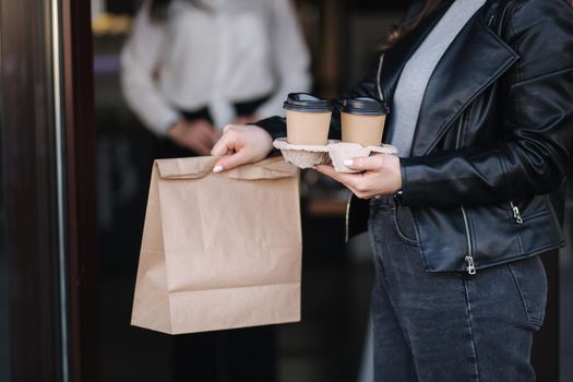 Attractive female customer in face mask standing outdoors by cafe and take away food and coffee. Background of cashier in mask waiting for next customer.