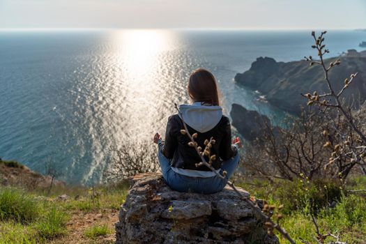 Woman tourist enjoying the sunset over the sea mountain landscape. Sits outdoors on a rock above the sea. She is wearing jeans, a blue hoodie and a black leather jacket. Healthy lifestyle, harmony and meditation.