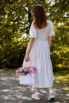 A beautiful bouquet of flowers in a box in the hands of a beautiful girl who walks along the street on a sunny day. Girl in a dress, glasses and sneakers. Focus on the background of flowers.