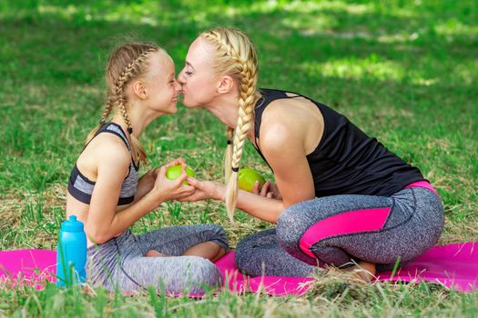 Mother kissing her daughter in nose giving green apple sitting on the grass in the park