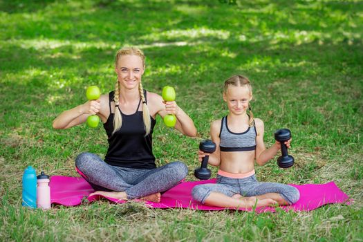 Young woman and girl are training with dumbbells sitting on the grass in the park