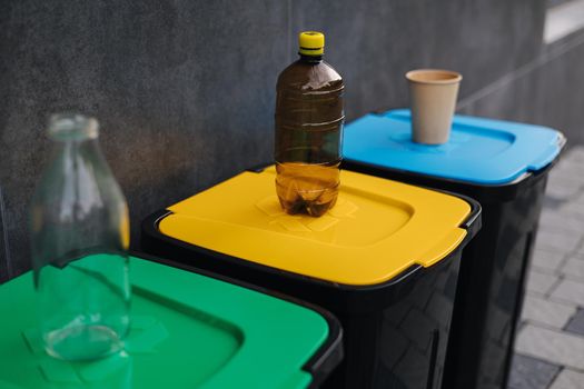 Outdoors in the park is recycling bins different colours. Tree closed plastic bins. Green, yeallow and blue. On top of each bin stand - glass bottle, plastic bottle and cardboard cup.