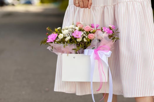 A beautiful bouquet of flowers in a box in the hands of a beautiful girl who walks along the street on a sunny day. Girl in a dress, glasses and sneakers. Focus on the background of flowers.