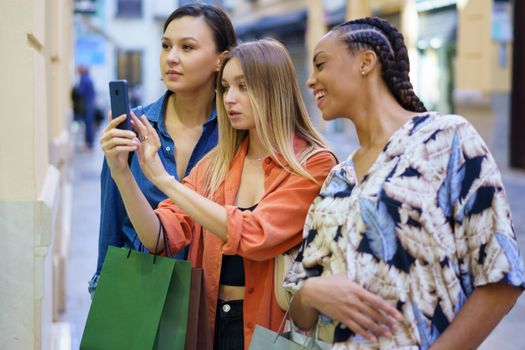 Group of diverse female friends with shopping bags taking picture of showcase on cellphone while choosing goods on street during shopping