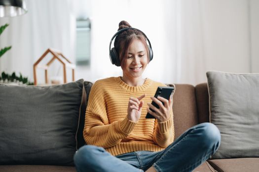 Freelance asian woman with mobile phone listening music in headphones and relax at home. Happy girl sitting on couch in living room