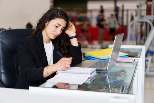 Young brunette in smart casual clothes touching hair and writing in notebook while sitting at desk with laptop during psychology session in gym