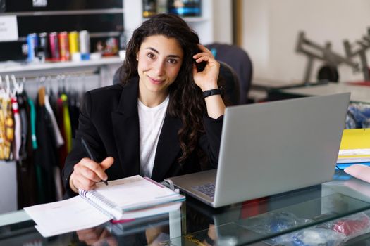 Positive young woman, in smart casual clothes, looking at camera with smile and touching dark hair, while sitting at desk and making notes during psychology session in gym