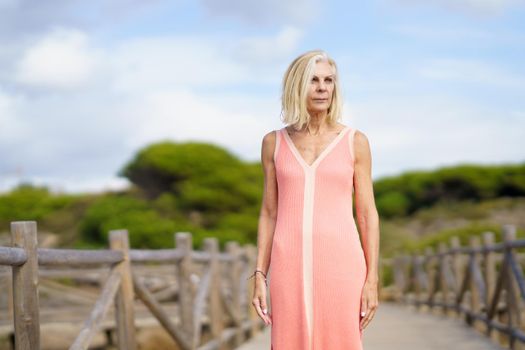 Beautiful mature woman walking along a wooden path near the beach., wearing a nice orange dress. Elderly female enjoying her retirement at a seaside retreat.