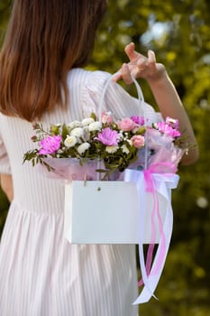 A beautiful bouquet of flowers in a box in the hands of a beautiful girl who walks along the street on a sunny day. Girl in a dress, glasses and sneakers. Focus on the background of flowers.