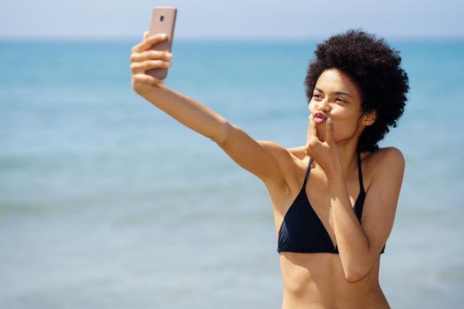 Cheerful African American female traveler in bikini pouting lips and taking self portrait on smartphone near sea in tropical resort