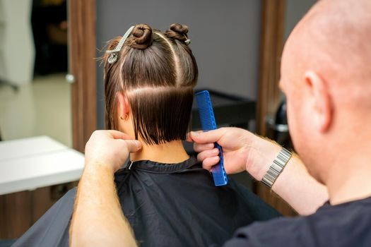 Male hairdresser cutting hair of young woman holding comb at hair salon. Rear view