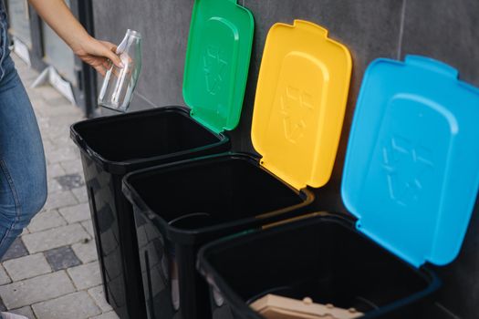 Close-up of female throwing glass bottle in recycling bin. Different colour of recycling bins outdoors. Side view of woman put rubbish in container.