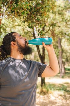 side image of a young fitness man drinking water from a bottle after a sports workout to refresh herself. boy practising sport outdoors. health and wellness lifestyle.outdoor public park, natural sunlight.vertical