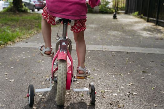 Closeup of girls' feet with white sandals on a pink bike with 4 wheels